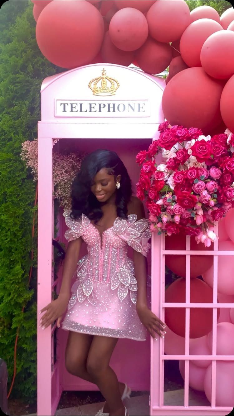 Young woman in pink dress posing in a pink telephone booth with balloons.