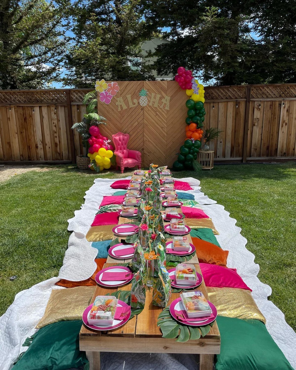 Tropical party table setup with pink plates, colorful cushions, and Aloha backdrop.