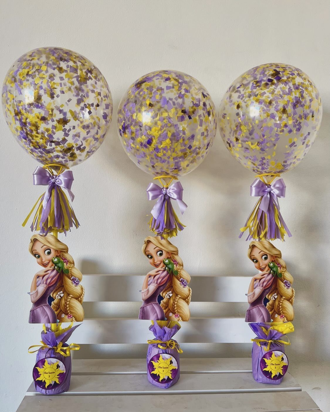 Three Tangled themed centerpieces with confetti balloons, Rapunzel cutouts, and purple gift bags.
