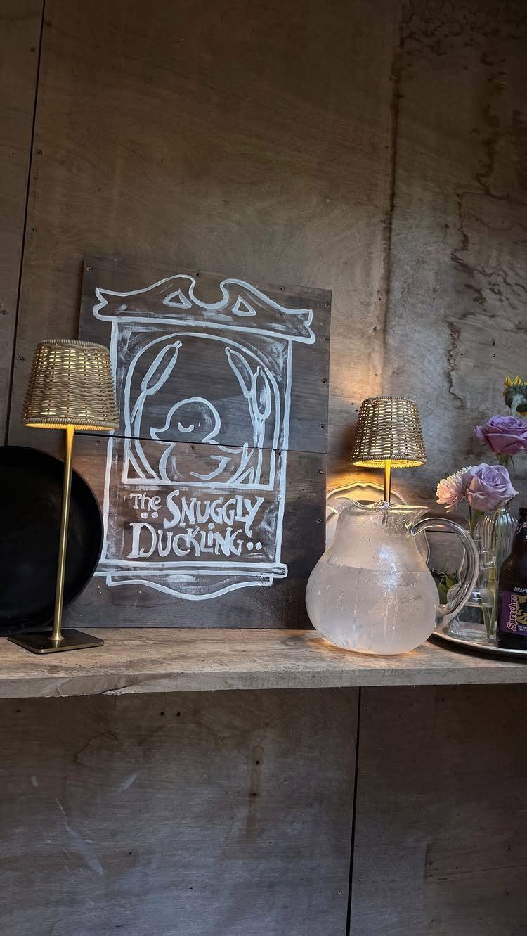 The Snuggly Duckling sign on a wooden shelf with lamps and water pitcher.
