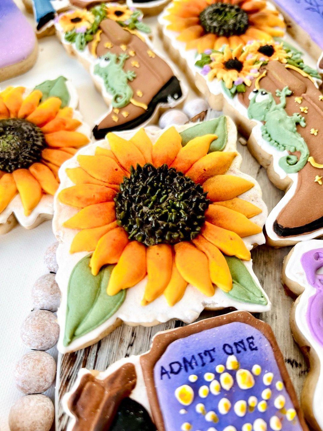 Decorated sugar cookies featuring sunflowers, brown boots, a green chameleon, and a purple ticket.