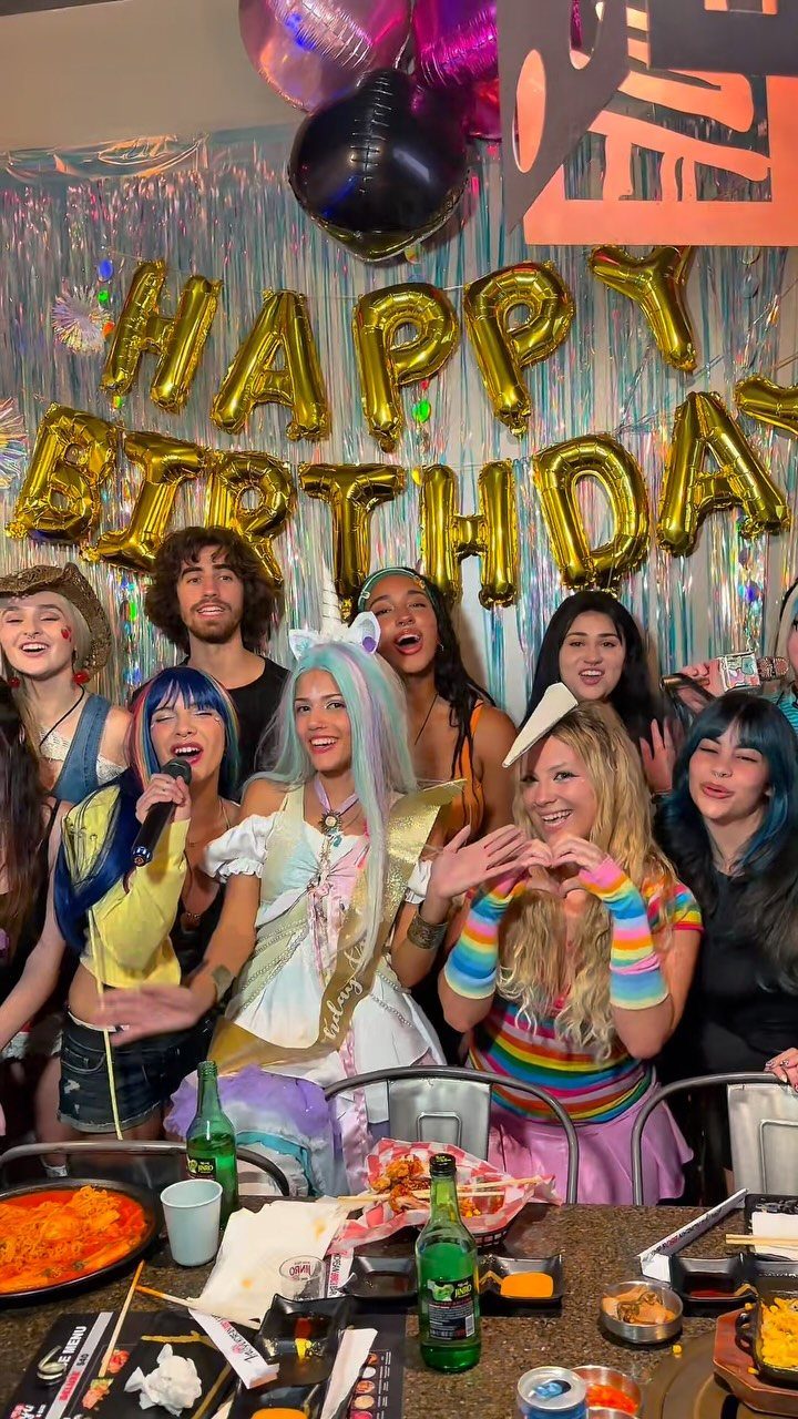 Teenagers in colorful costumes posing before a gold Happy Birthday balloon backdrop.