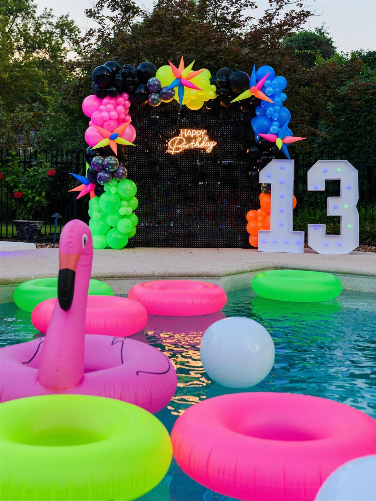 Swimming pool with neon inflatable floats and a colorful birthday balloon backdrop.