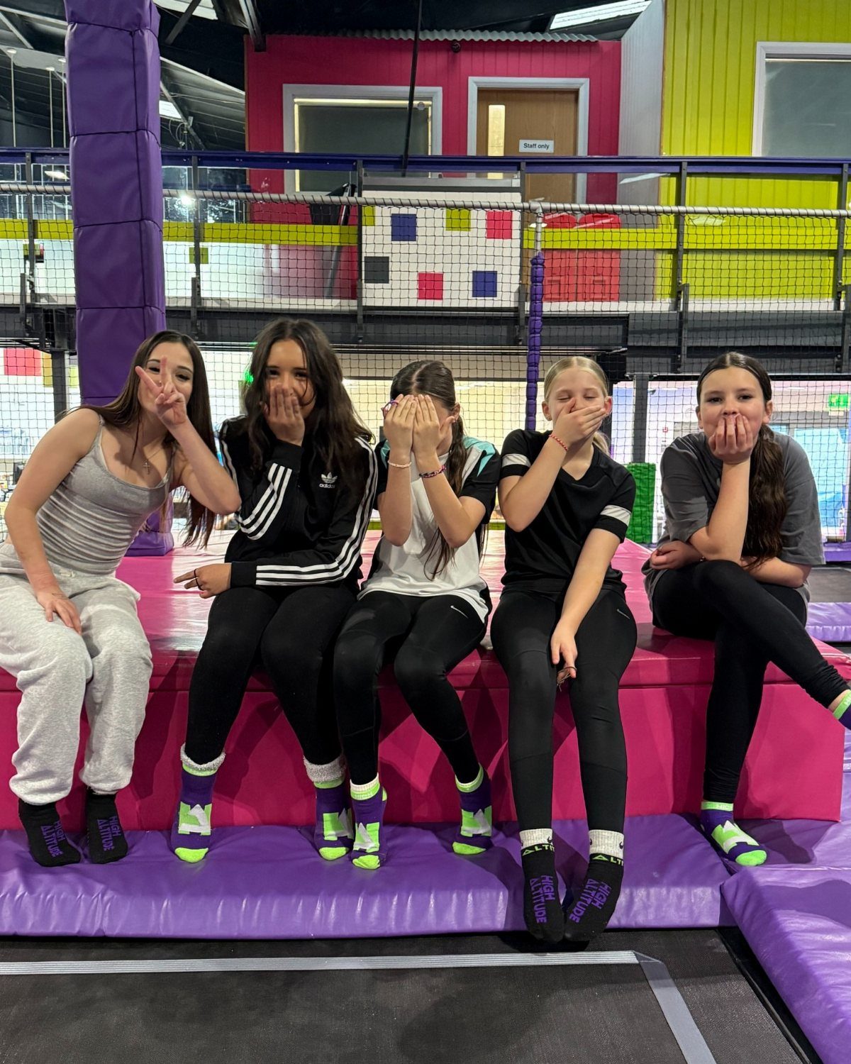 Five teenage girls sitting on pink padding inside a colorful trampoline park.