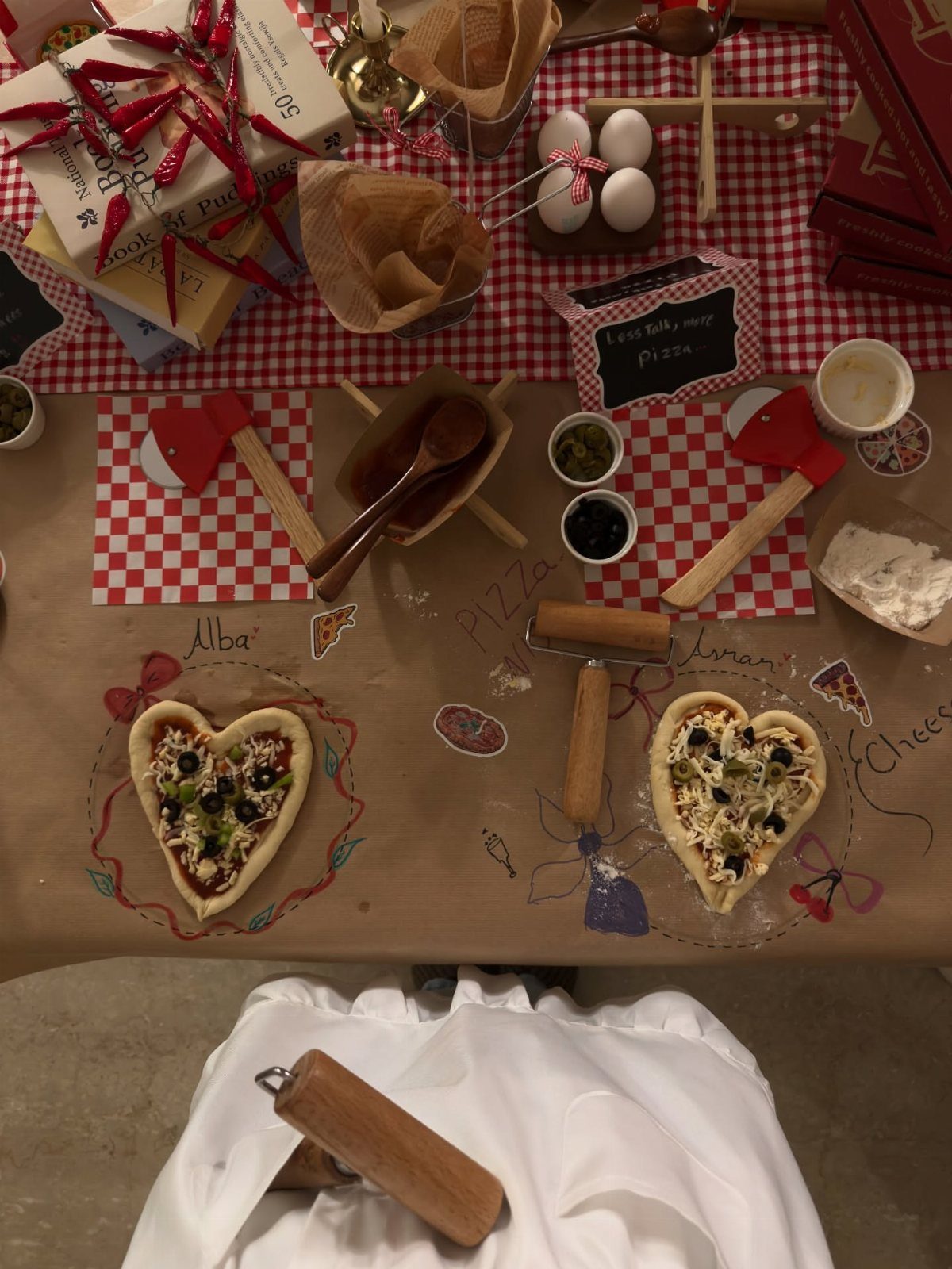 Two heart-shaped pizzas with toppings on a table with rolling pins and ingredients.