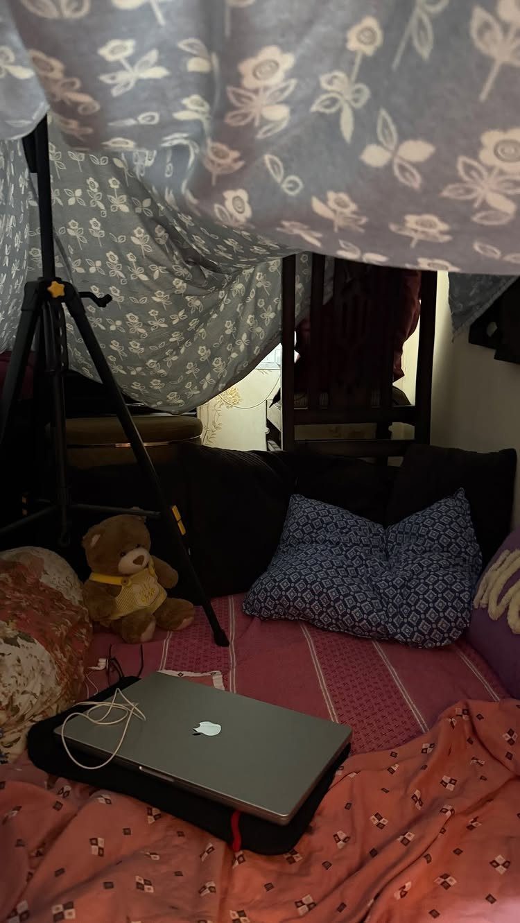Cozy pillow fort with floral sheets, pillows, a teddy bear, and a laptop.