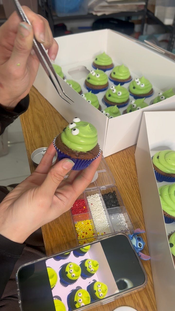 Green alien cupcakes being decorated with small white eyes using metal tweezers.
