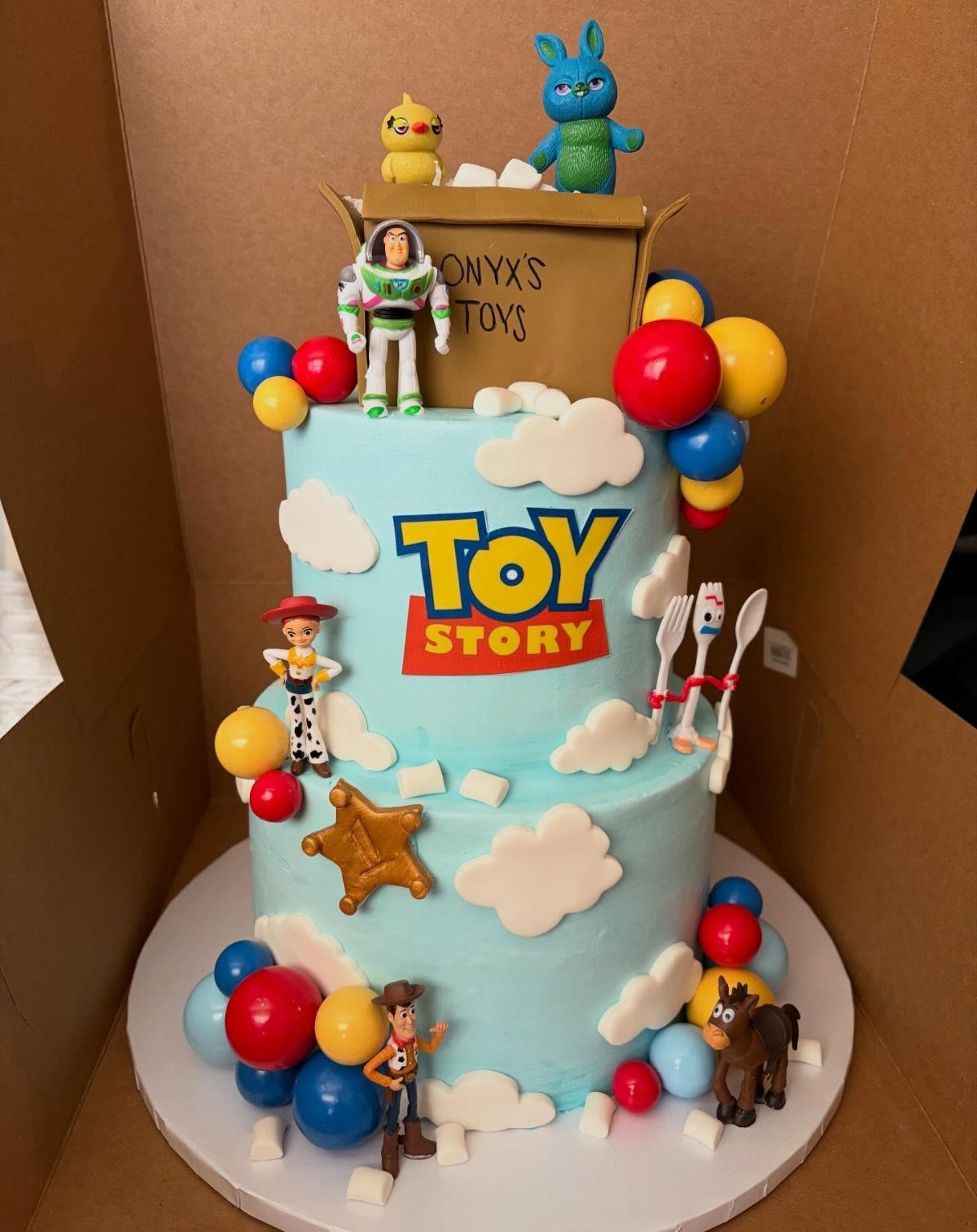 Three-tier blue Toy Story cake with character figures, clouds, and colorful candy decorations.