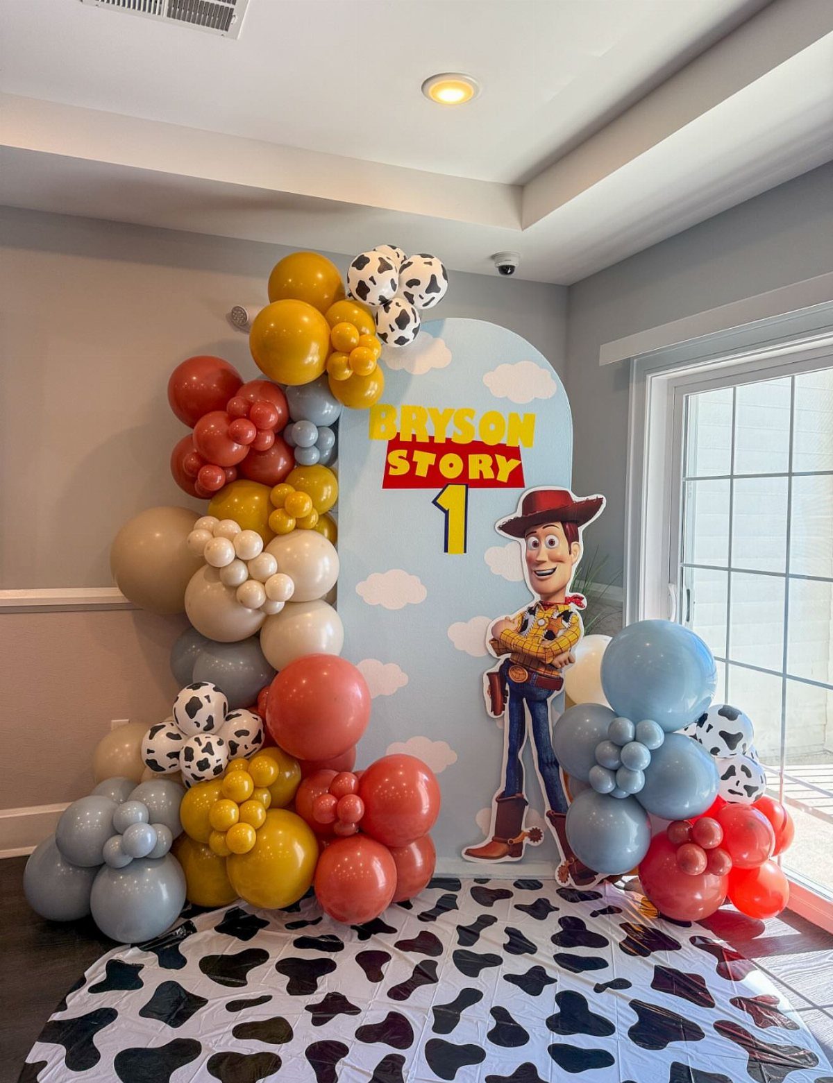 Toy Story party backdrop with Woody cutout, colorful balloons, and cow-print floor mat.