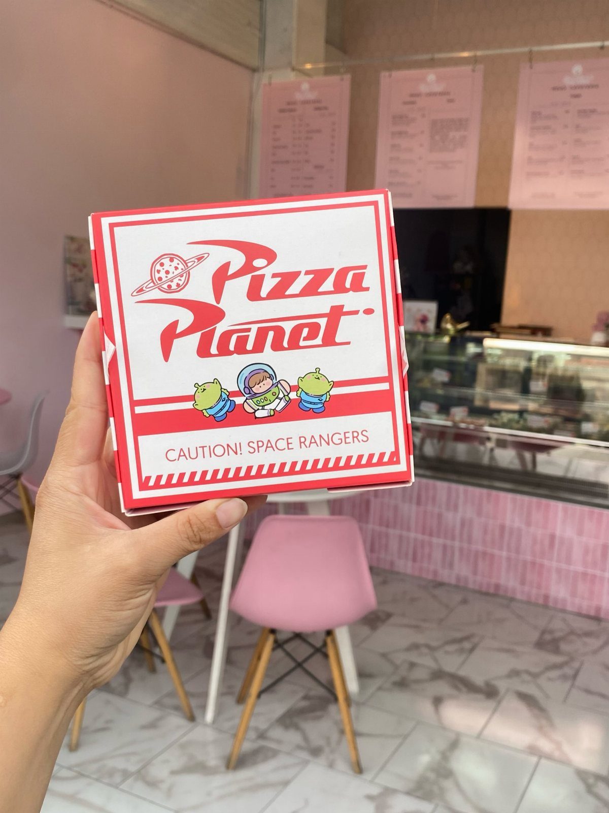 Pizza Planet pizza box held by a hand in a pink themed room.