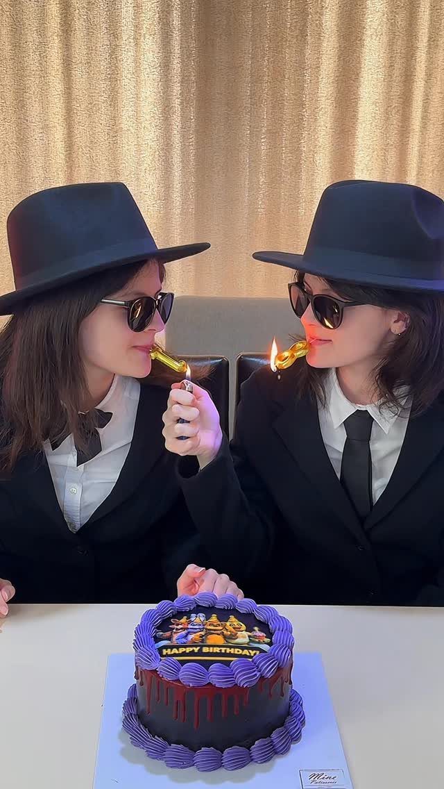 Two women in matching black suits and fedoras lighting a birthday cake.