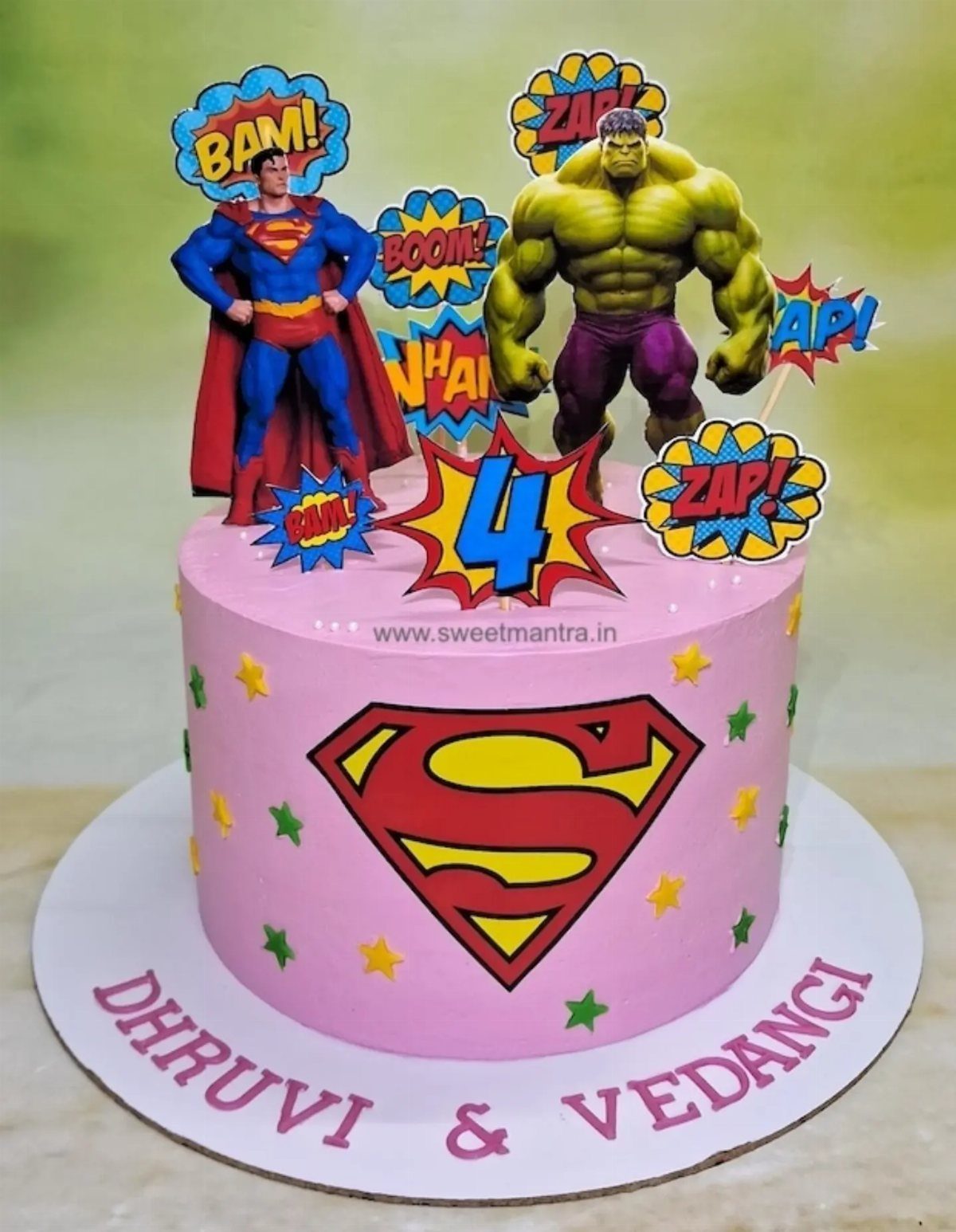 Pink birthday cake with Superman and Hulk figurines and comic book speech bubbles.