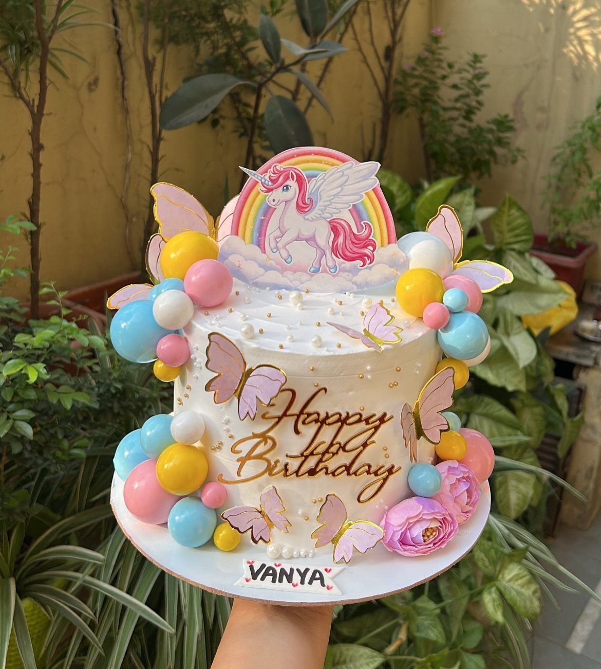 White birthday cake with pastel balloons, pink butterflies, and a unicorn rainbow topper.