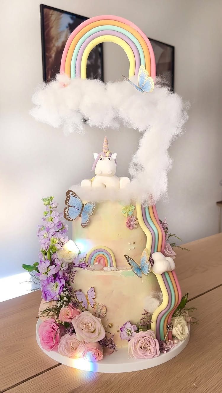 Multi-tiered white cake with a rainbow arch, unicorn figurine, and colorful floral decorations.