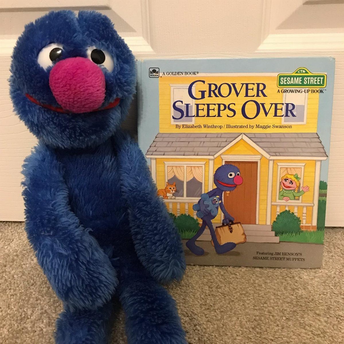 Grover plush toy sitting beside a Grover Sleeps Over children's book on carpet.